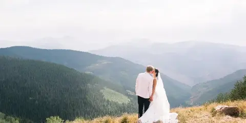 A bride and groom standing on a grassy mountain ridge overlooking a vast valley and forested peaks.