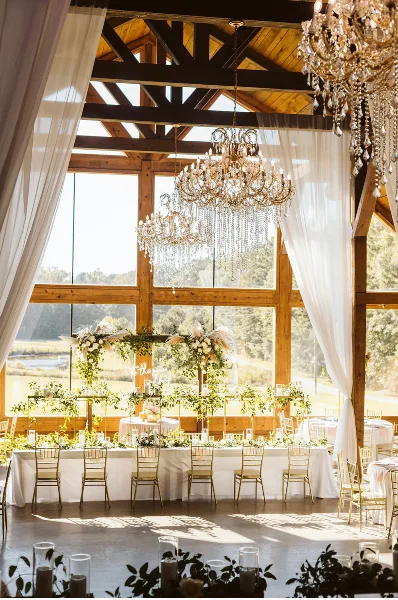 Interior view of a rustic barn wedding venue featuring vaulted wooden ceilings, crystal chandeliers, and a large window wall overlooking a grassy field.