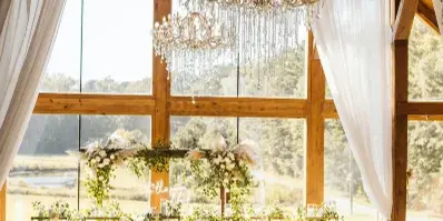 Interior view of a rustic barn wedding venue featuring vaulted wooden ceilings, crystal chandeliers, and a large window wall overlooking a grassy field.