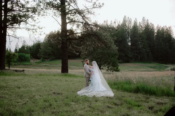 A bride and groom standing in a grassy field with a backdrop of tall evergreen trees.