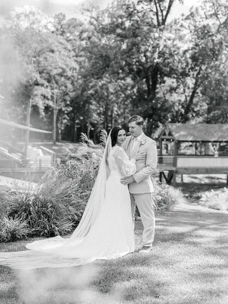 A black-and-white photograph of a bride and groom standing together outdoors, with the bride in a long gown and veil and the groom in a light suit.