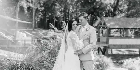 A black-and-white photograph of a bride and groom standing together outdoors, with the bride in a long gown and veil and the groom in a light suit.