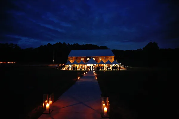 A wedding ceremony setup at dusk featuring a long aisle lined with candles leading to a white tent and a large event barn.