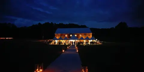 A wedding ceremony setup at dusk featuring a long aisle lined with candles leading to a white tent and a large event barn.