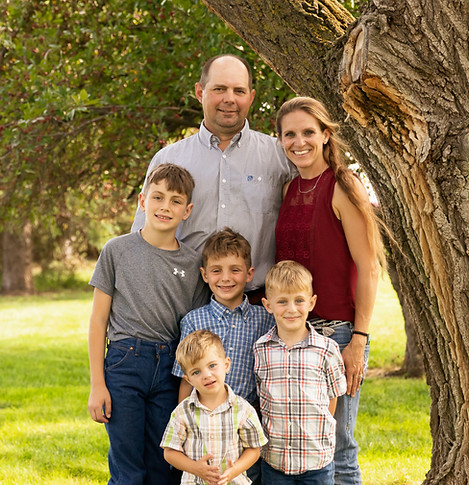 A professional outdoor family portrait of a man, a woman, and four young boys standing together in a grassy area next to a large tree.