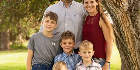A professional outdoor family portrait of a man, a woman, and four young boys standing together in a grassy area next to a large tree.