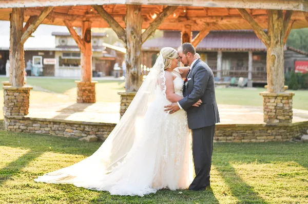 A bride and groom embracing in front of a rustic wooden pavilion with stone pillars at a farm venue.