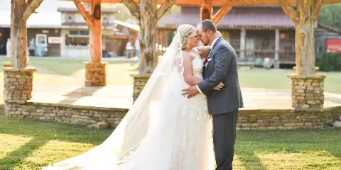 A bride and groom embracing in front of a rustic wooden pavilion with stone pillars at a farm venue.