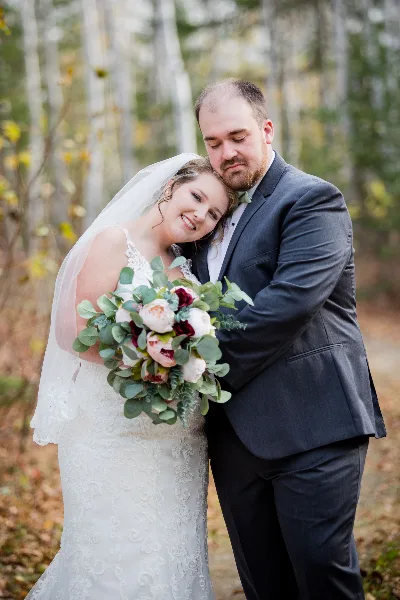 A bride and groom embracing in a forest setting, with the bride holding a bouquet of white and burgundy flowers.