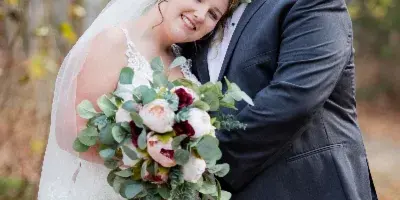 A bride and groom embracing in a forest setting, with the bride holding a bouquet of white and burgundy flowers.