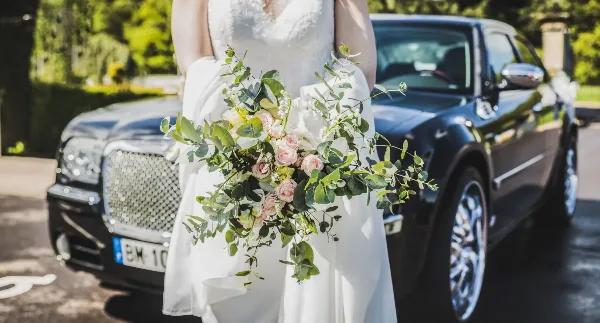 A bride in a white wedding dress holding a floral bouquet standing in front of a black luxury sedan.