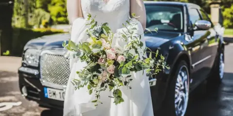A bride in a white wedding dress holding a floral bouquet standing in front of a black luxury sedan.