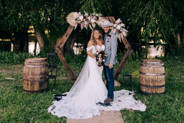 A bride and groom standing under a rustic wooden arch decorated with flowers and pampas grass, flanked by wooden barrels.