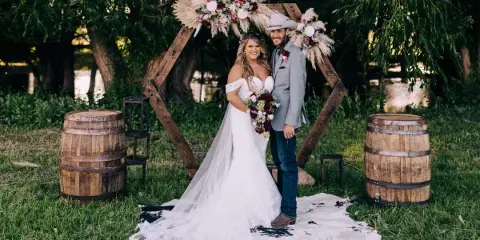 A bride and groom standing under a rustic wooden arch decorated with flowers and pampas grass, flanked by wooden barrels.