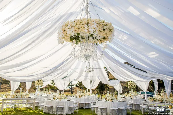 Outdoor wedding reception setup featuring a large white tent with draped fabric, a hanging floral chandelier, and round tables set with white linens and chairs.