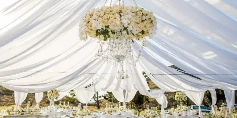 Outdoor wedding reception setup featuring a large white tent with draped fabric, a hanging floral chandelier, and round tables set with white linens and chairs.
