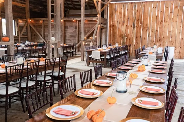 A rustic wedding reception setup with long wooden farm tables, dark Chiavari chairs, and table settings.