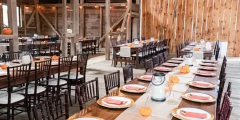 A rustic wedding reception setup with long wooden farm tables, dark Chiavari chairs, and table settings.