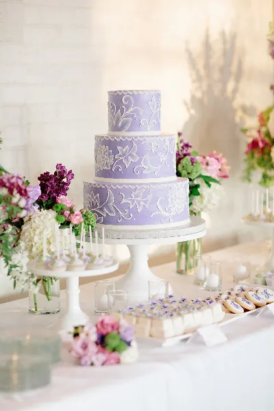 A three-tiered lavender wedding cake with white floral piping on a white stand, surrounded by flowers and a dessert bar.