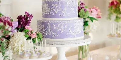 A three-tiered lavender wedding cake with white floral piping on a white stand, surrounded by flowers and a dessert bar.