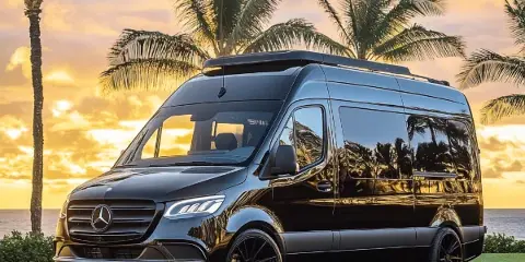 A black Mercedes-Benz Sprinter van parked on a paved surface with palm trees and a sunset in the background.