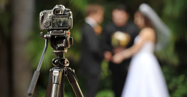 A professional video camera on a tripod in the foreground, focused on a wedding couple exchanging vows in the blurred background.