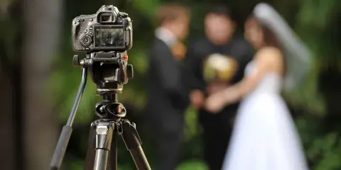 A professional video camera on a tripod in the foreground, focused on a wedding couple exchanging vows in the blurred background.