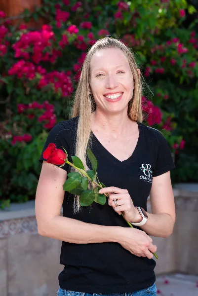 A smiling woman in a black Bella Rose Estate t-shirt holding a red rose in front of a garden wall with pink flowers.