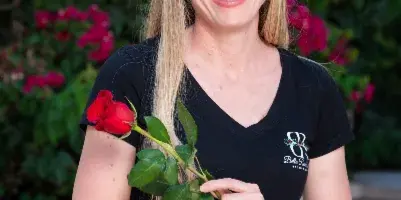 A smiling woman in a black Bella Rose Estate t-shirt holding a red rose in front of a garden wall with pink flowers.