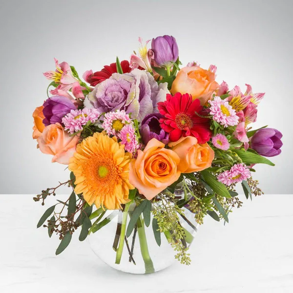 A colorful mixed bouquet of roses, gerbera daisies, tulips, and chrysanthemums arranged in a clear glass vase.
