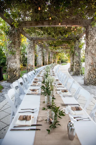 A long outdoor wedding table set with white linens, greenery, and floral arrangements under a vine-covered pergola.