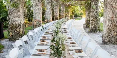 A long outdoor wedding table set with white linens, greenery, and floral arrangements under a vine-covered pergola.