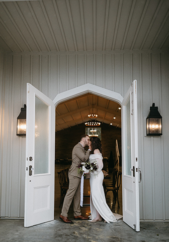 A couple kissing in front of the white double entry doors leading into the Great Hall of The Woods at Cedar Springs wedding venue.