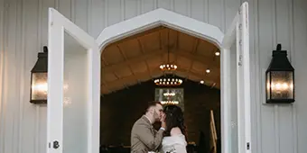 A couple kissing in front of the white double entry doors leading into the Great Hall of The Woods at Cedar Springs wedding venue.