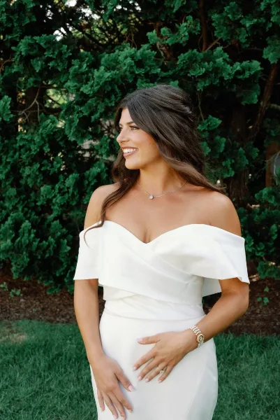 Bride in an off-the-shoulder white gown with styled hair and makeup standing outdoors in front of greenery.