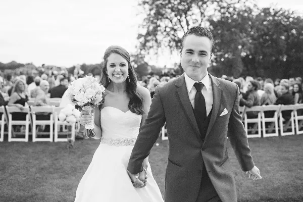 A black and white photograph of a bride and groom walking hand-in-hand down an outdoor wedding aisle.