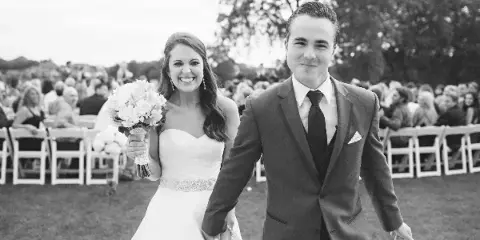 A black and white photograph of a bride and groom walking hand-in-hand down an outdoor wedding aisle.