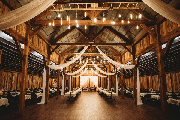 Interior of a rustic timber-framed barn wedding venue featuring exposed wooden beams, hanging string lights, and a dance floor.