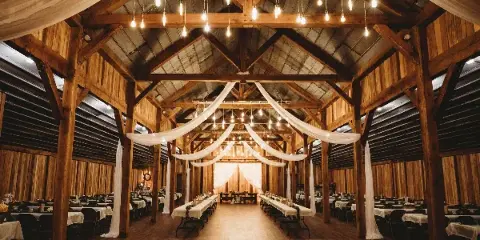 Interior of a rustic timber-framed barn wedding venue featuring exposed wooden beams, hanging string lights, and a dance floor.