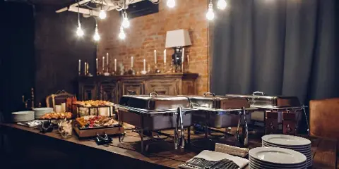 A catering buffet table set with stainless steel chafing dishes, plates, and food trays in an event space.