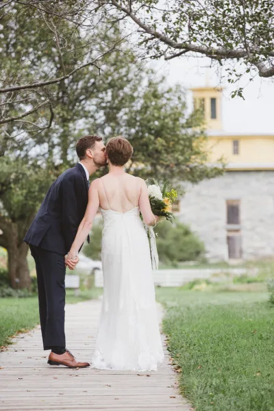 A bride and groom kissing on a wooden boardwalk with a stone building and trees in the background.