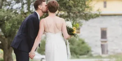 A bride and groom kissing on a wooden boardwalk with a stone building and trees in the background.
