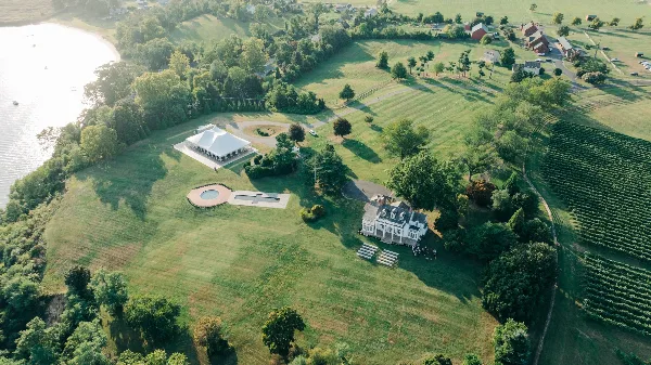 Aerial view of the Bohemia Overlook wedding venue estate featuring a white event tent, a historic-style building, and a vineyard adjacent to a river.