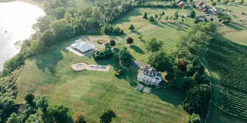 Aerial view of the Bohemia Overlook wedding venue estate featuring a white event tent, a historic-style building, and a vineyard adjacent to a river.