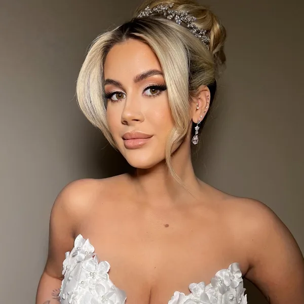 A woman with an elegant updo hairstyle, diamond headpiece, and full bridal makeup wearing a white floral wedding dress.