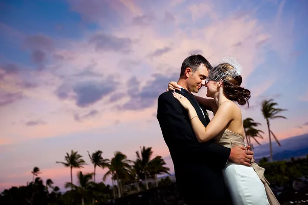 A bride and groom embracing in a romantic pose against a sunset sky with palm trees in the background.