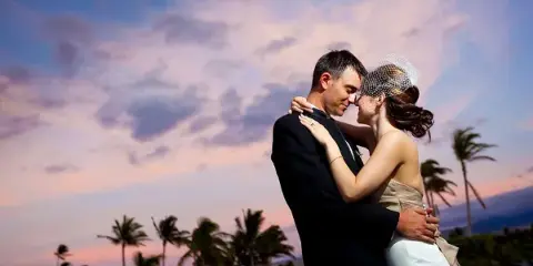 A bride and groom embracing in a romantic pose against a sunset sky with palm trees in the background.