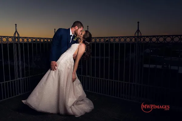 A bride and groom sharing a kiss on a rooftop terrace at dusk, wearing formal wedding attire.