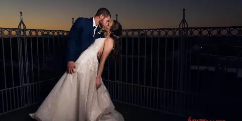A bride and groom sharing a kiss on a rooftop terrace at dusk, wearing formal wedding attire.