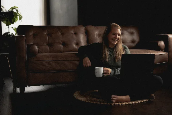 A female photographer sitting on the floor of a studio with a laptop and a mug, in front of a brown leather sofa.